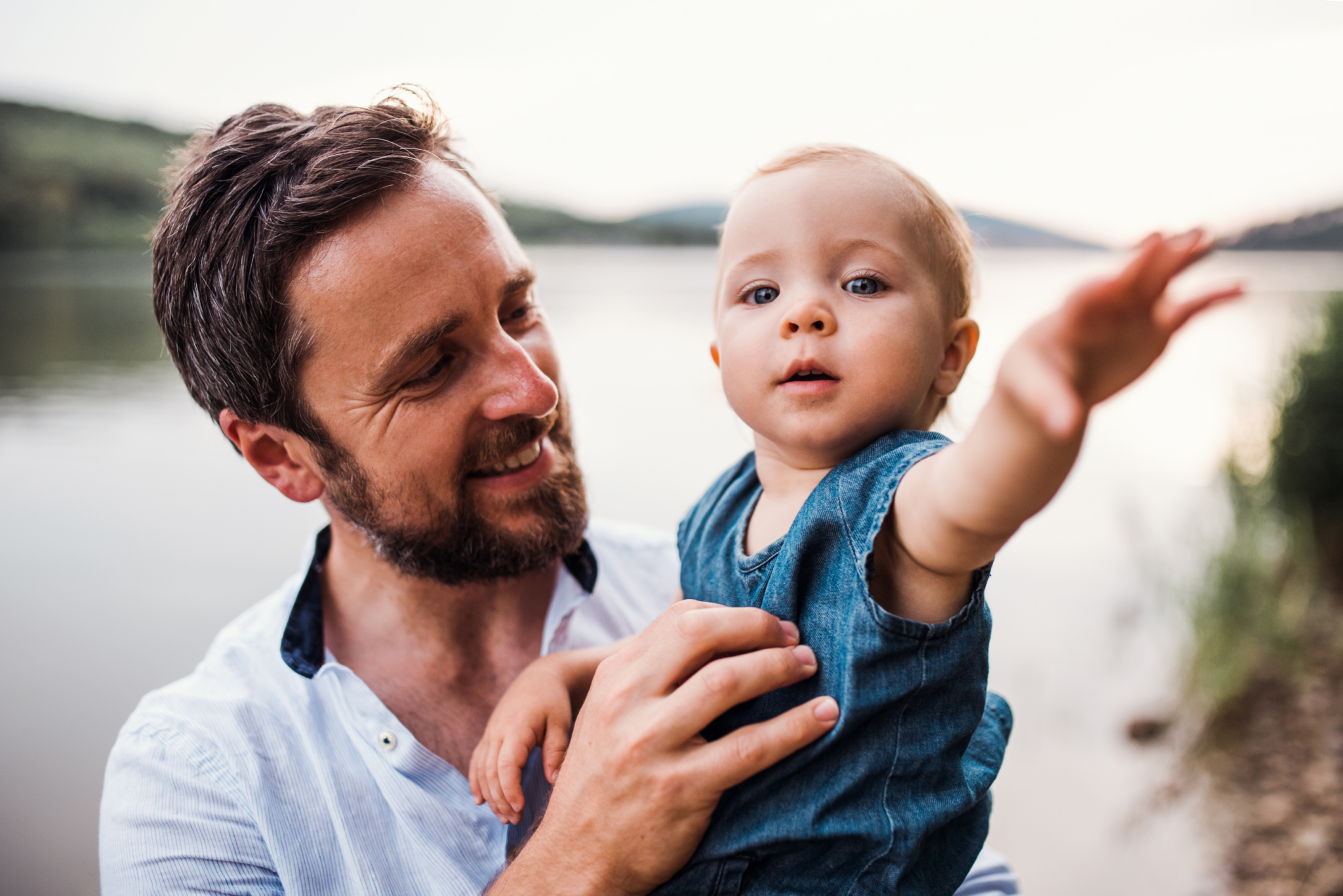 A man holding a baby by a lake.