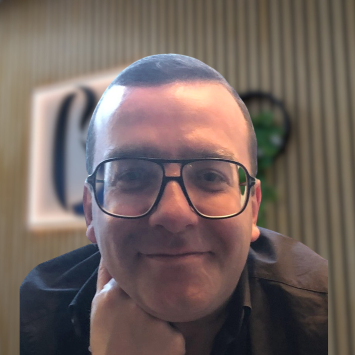 A person with short hair and glasses, smiling, rests their chin on their hand. In the background, a blurred wall with vertical lines and a framed object adds depth—capturing a candid moment reminiscent of Vincenzo Zuccarello’s relaxed and approachable style. Body Unity