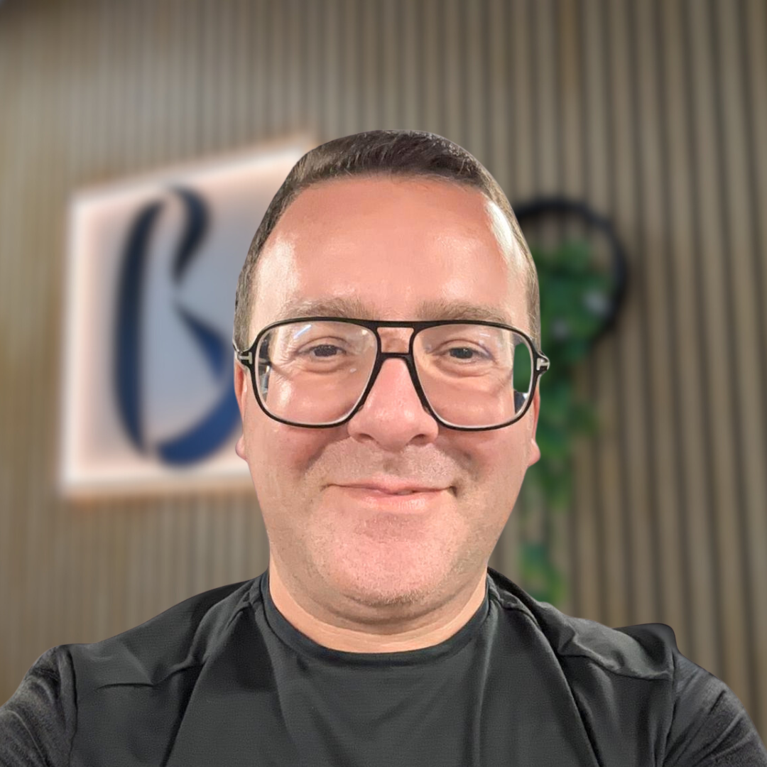 A person with short brown hair and glasses smiles at the camera, wearing a black shirt. The background is blurred, showing a sign with a blue letter and a plant. Body Unity